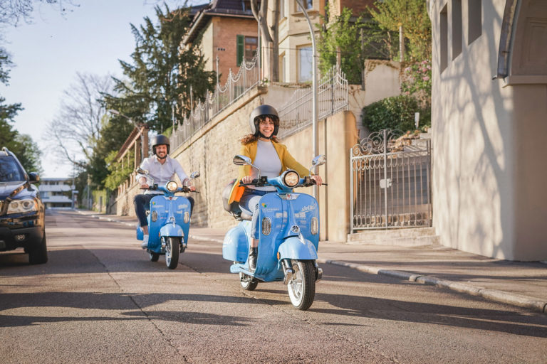 Ein Mann und eine Frau fahren jeweils auf einem Roller eine Straße entlang